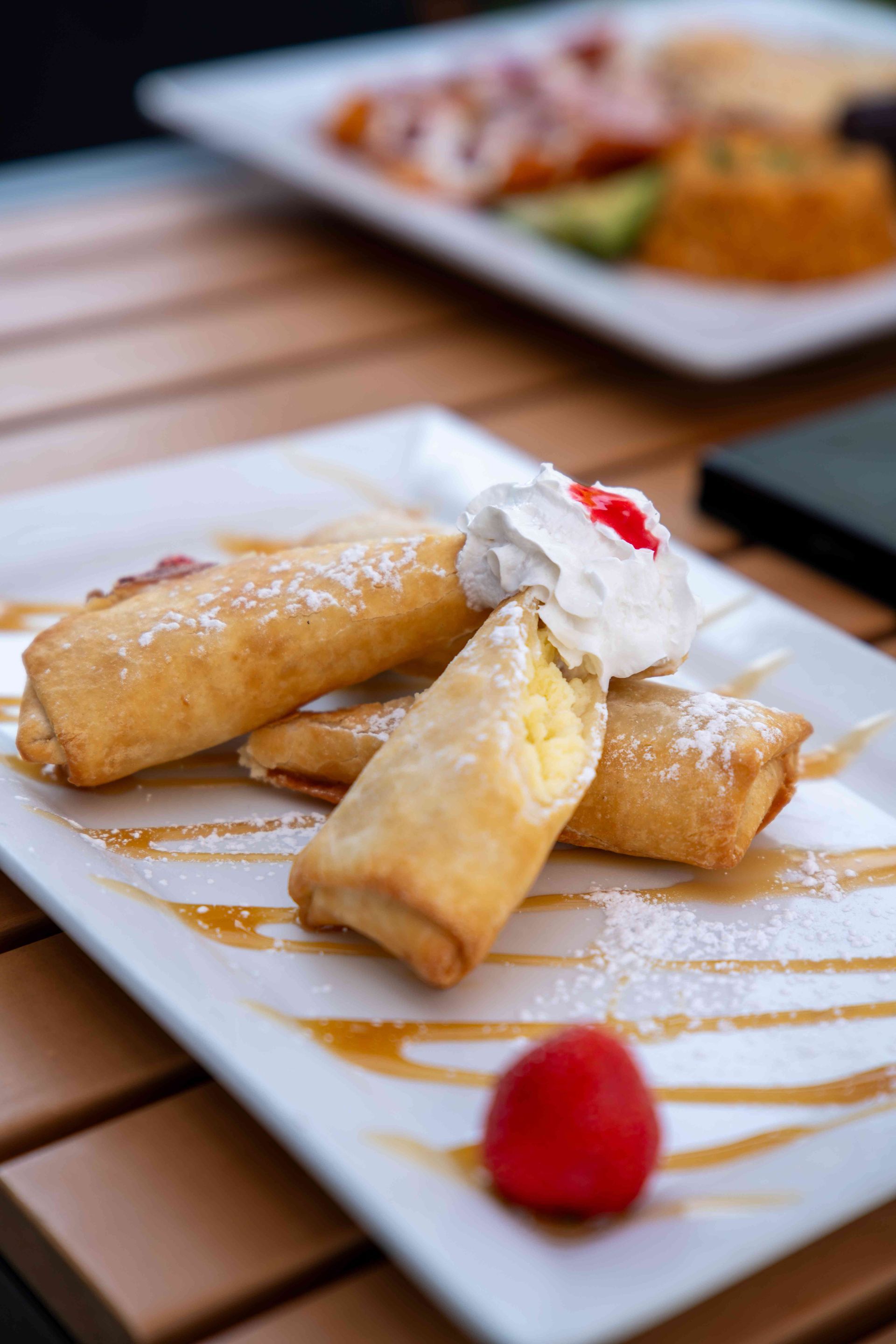 Fried dessert rolls on a white plate, drizzled with caramel and dusted with powdered sugar, topped with whipped cream and a cherry.