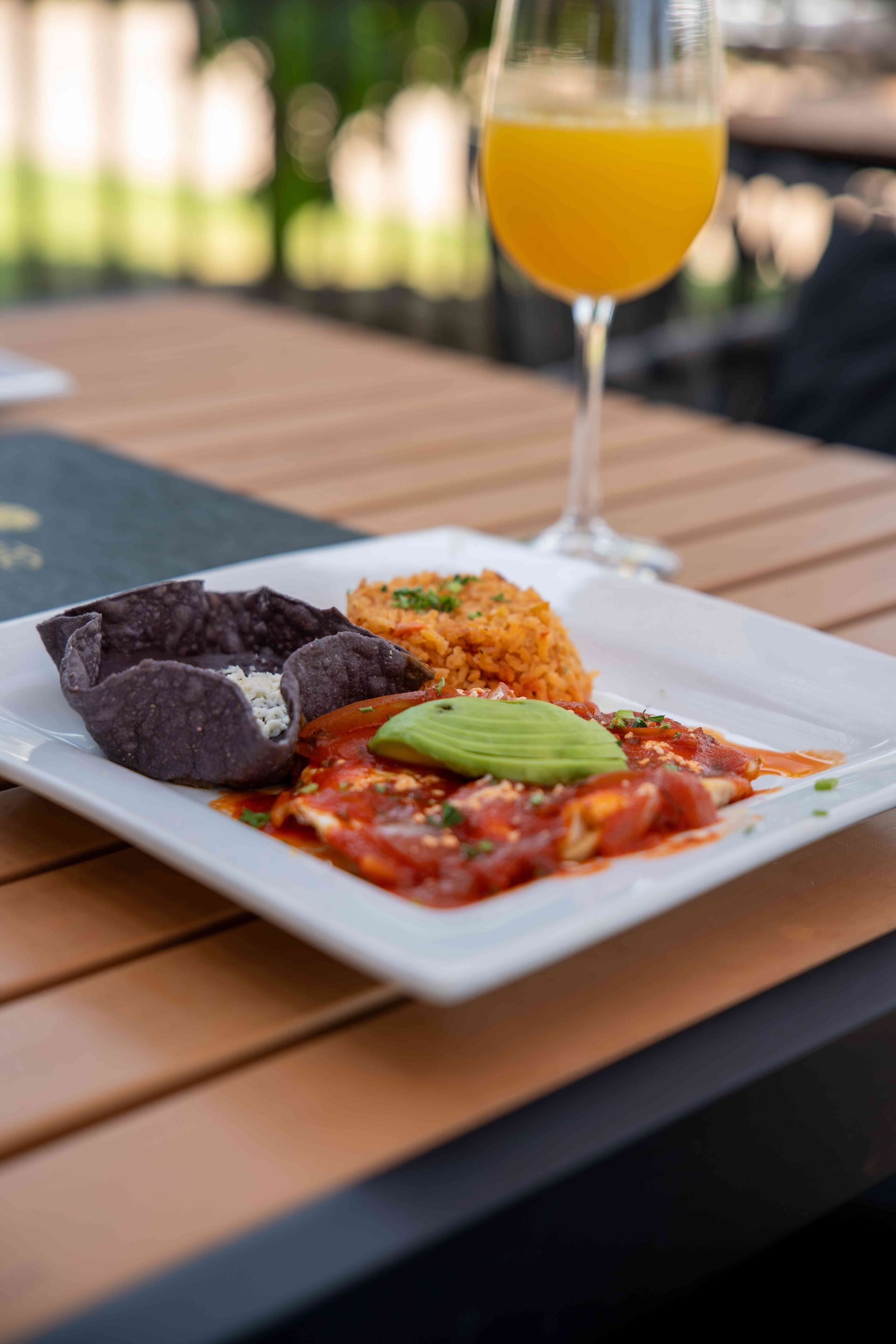 Breakfast plate with enchiladas, rice, avocado, and a blue corn tortilla. Mimosa in the background.