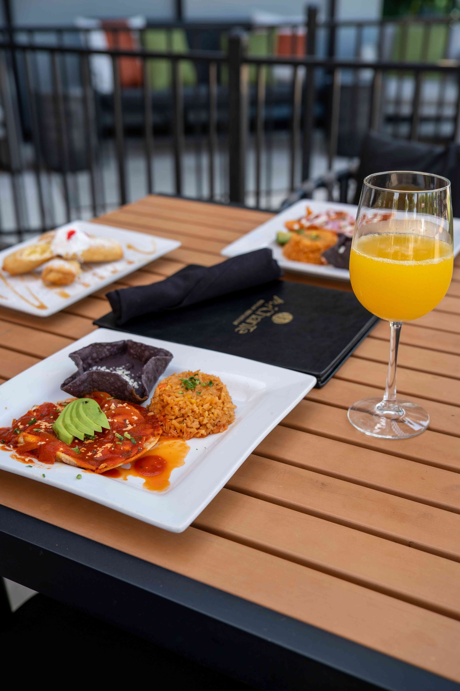 Mexican food and mimosa on a restaurant patio table; dishes of enchiladas, rice, pastries, and a drink.