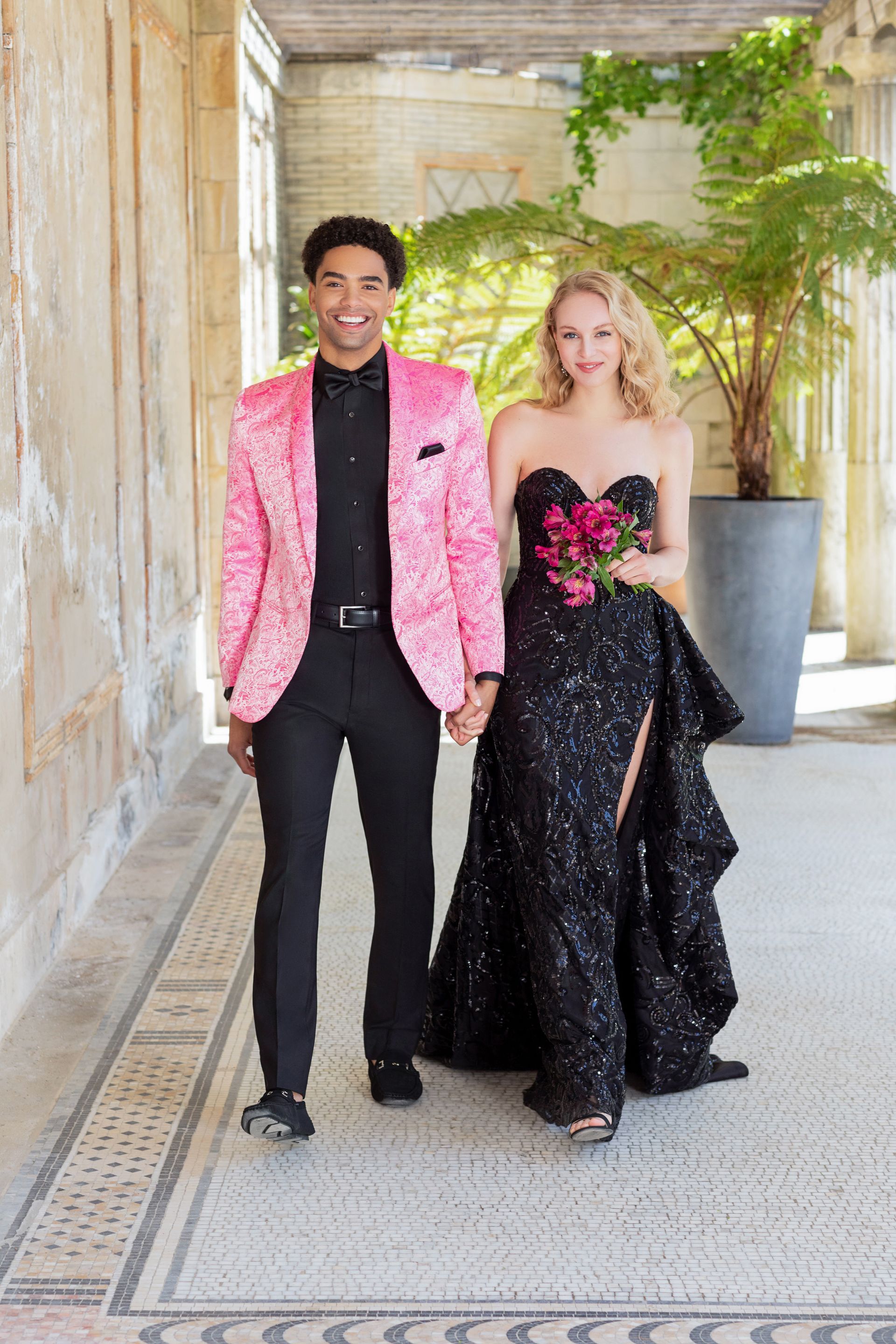 A person in a pink velvet blazer and another in a black sparkling strapless gown walk together in a tiled hallway.