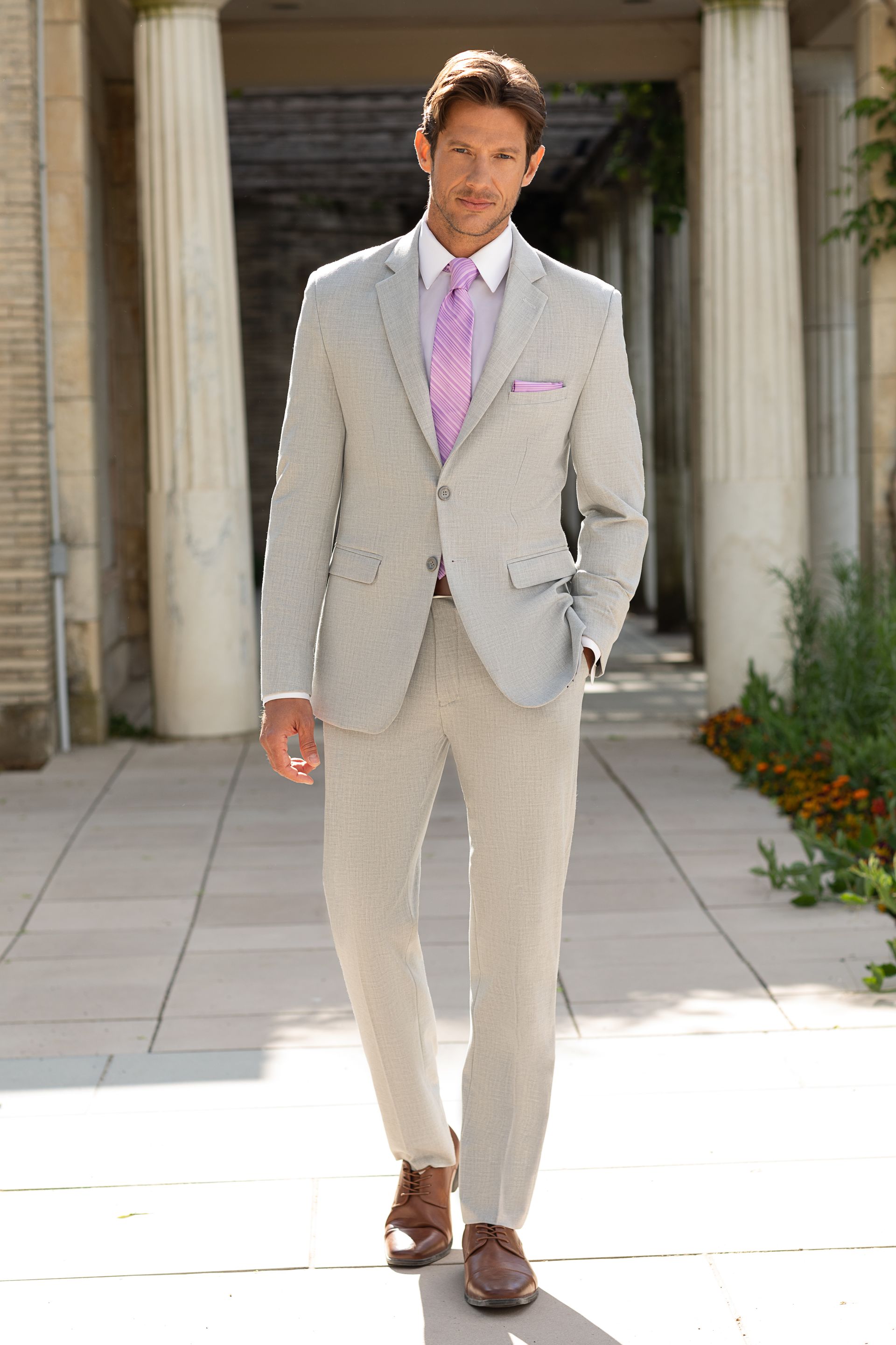 A man in a light grey suit and lavender tie walks outdoors in front of stone columns.