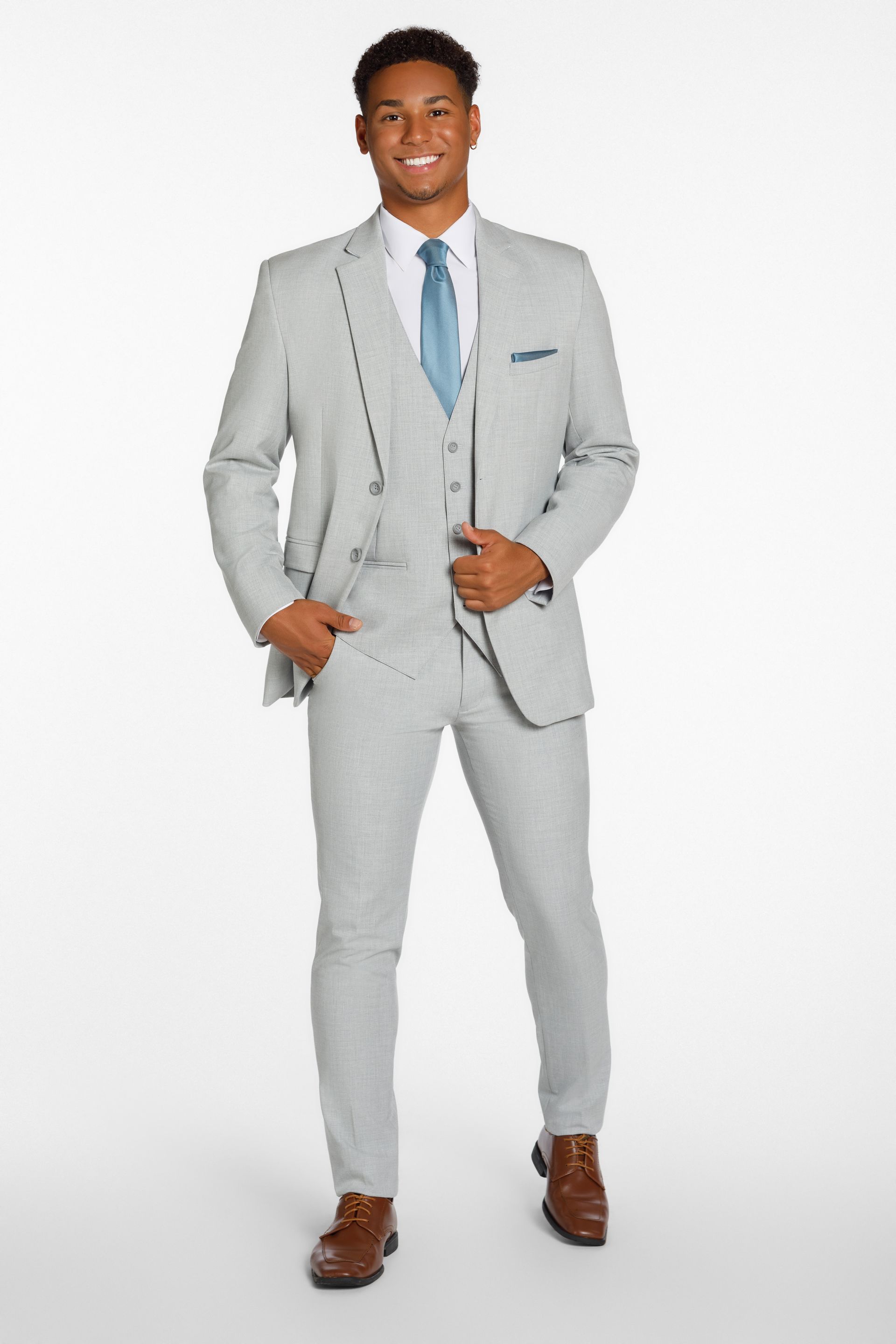 A smiling person stands wearing a light grey three-piece suit, white shirt, light blue tie, and brown dress shoes.