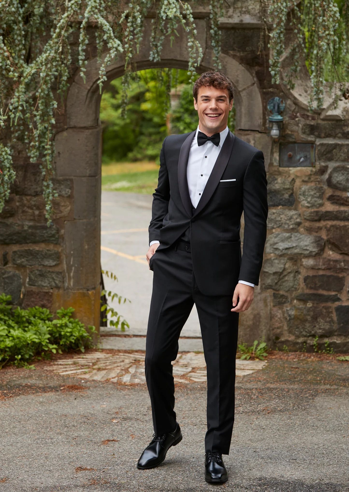 A smiling person in a black tuxedo stands in front of a stone archway with hanging greenery.