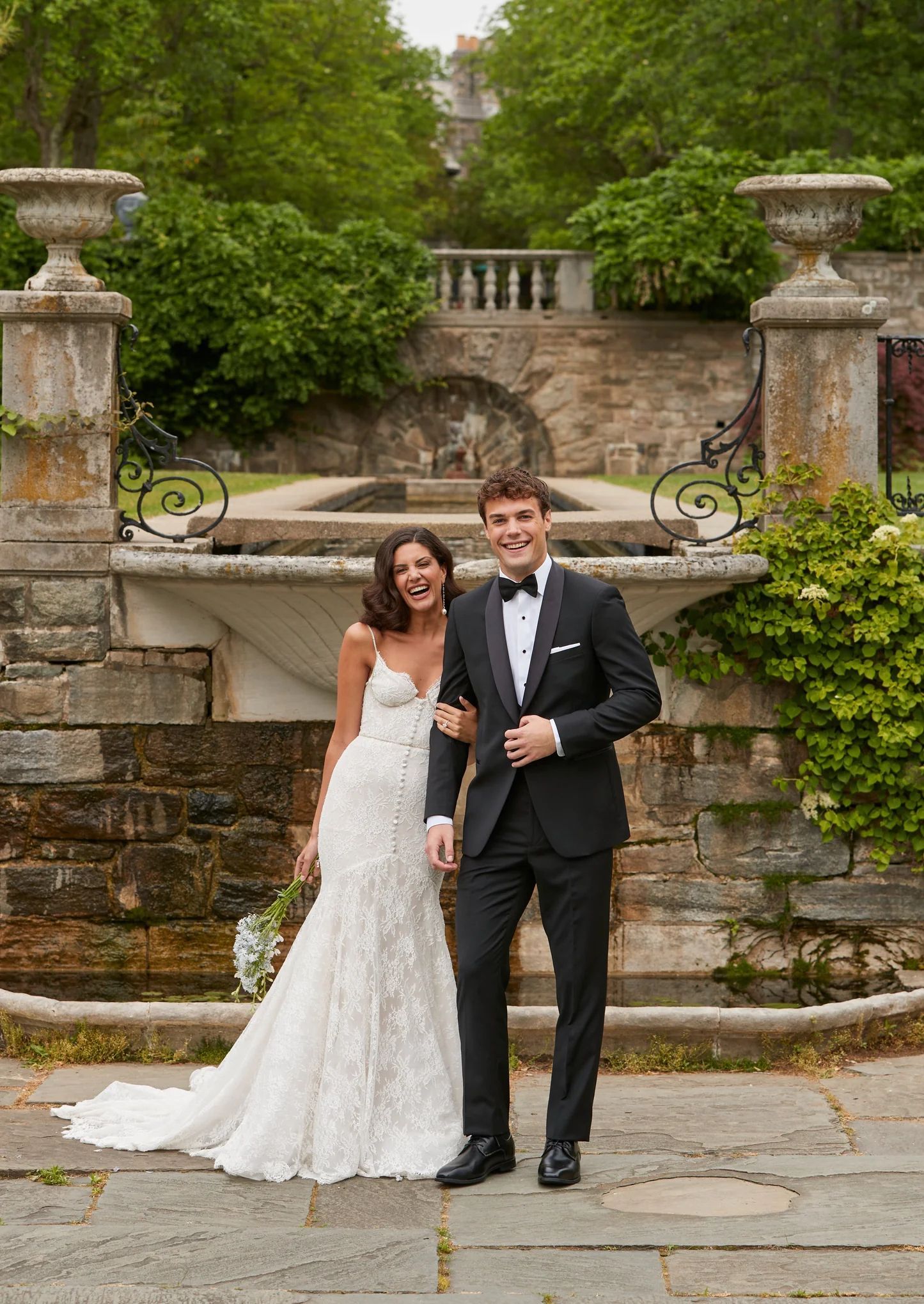 A smiling couple in formal wedding attire pose together outdoors in front of a stone fountain and garden.