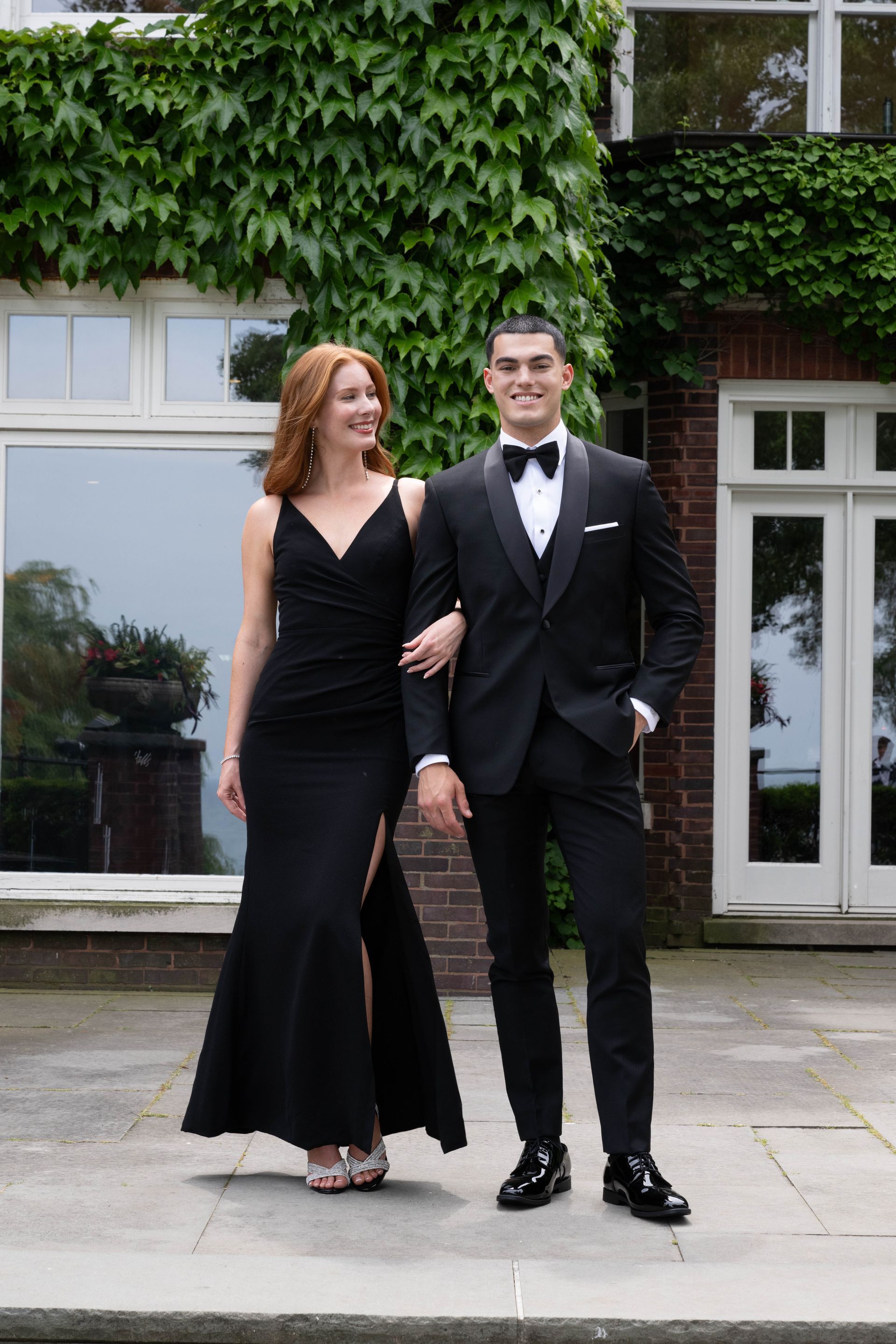 A couple dressed in formal black attire stands arm-in-arm on a stone patio in front of a building covered in ivy.