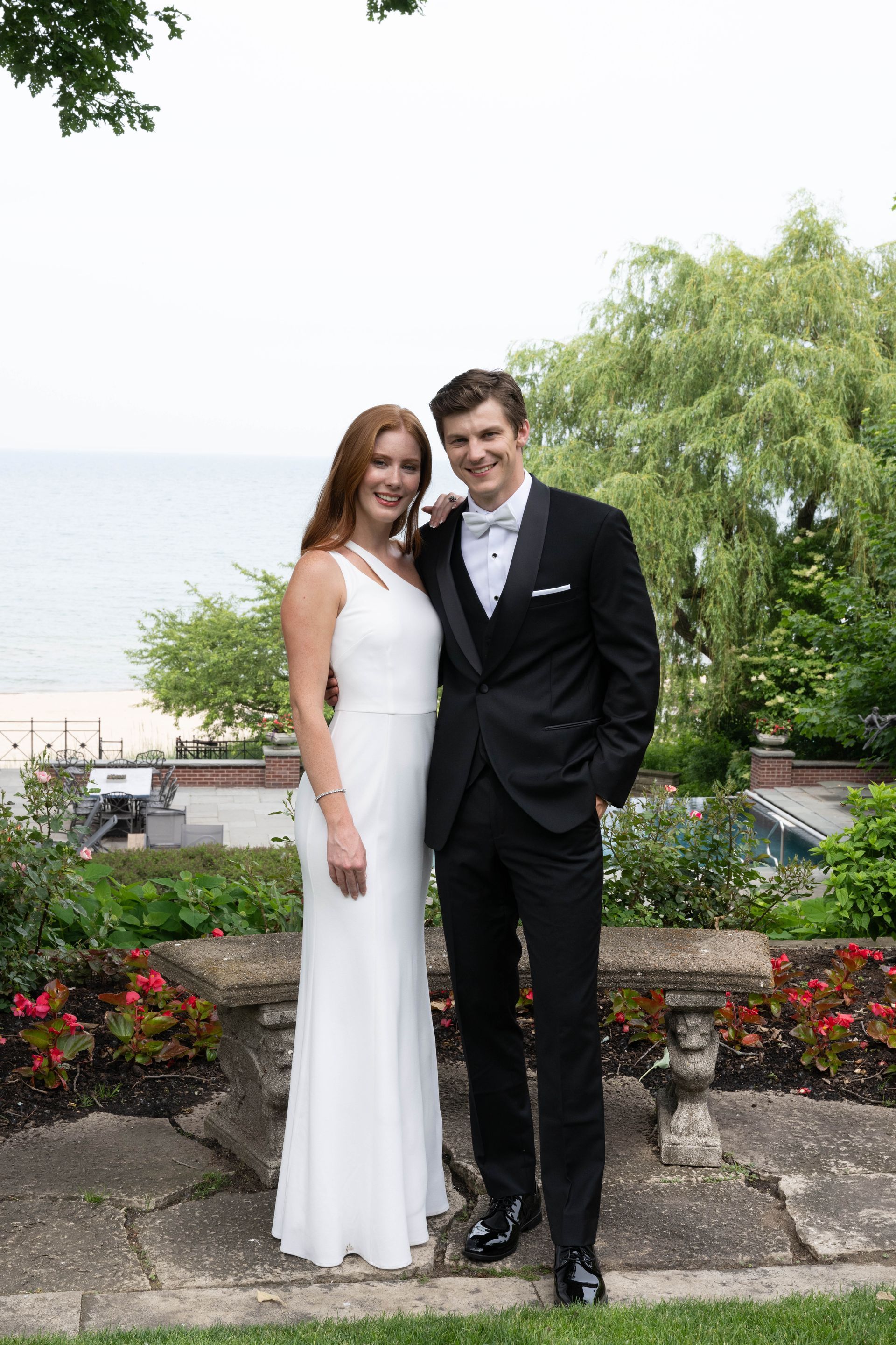 A smiling couple in formal wear stands together outdoors near a stone bench, with a waterfront backdrop and greenery.