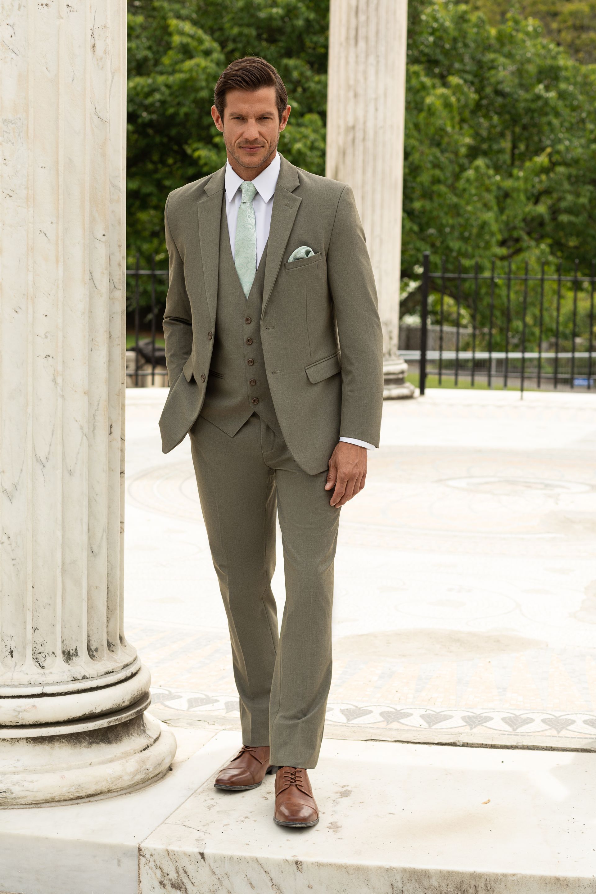 A person wearing a light green three-piece suit, white shirt, and mint tie, standing between marble columns outdoors.