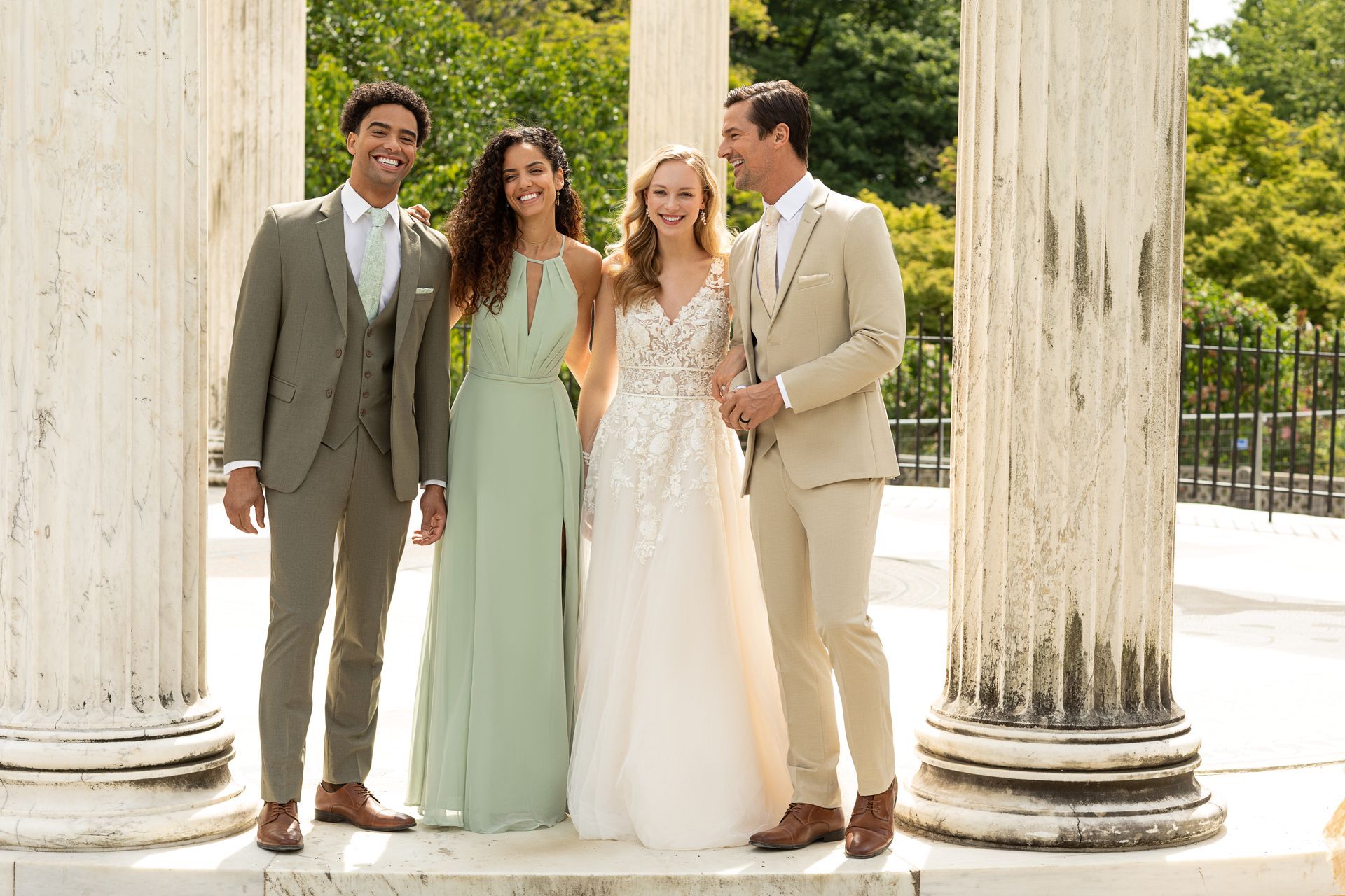 Two couples in formal wedding attire stand together outdoors between white stone pillars, smiling and posing for a photo.