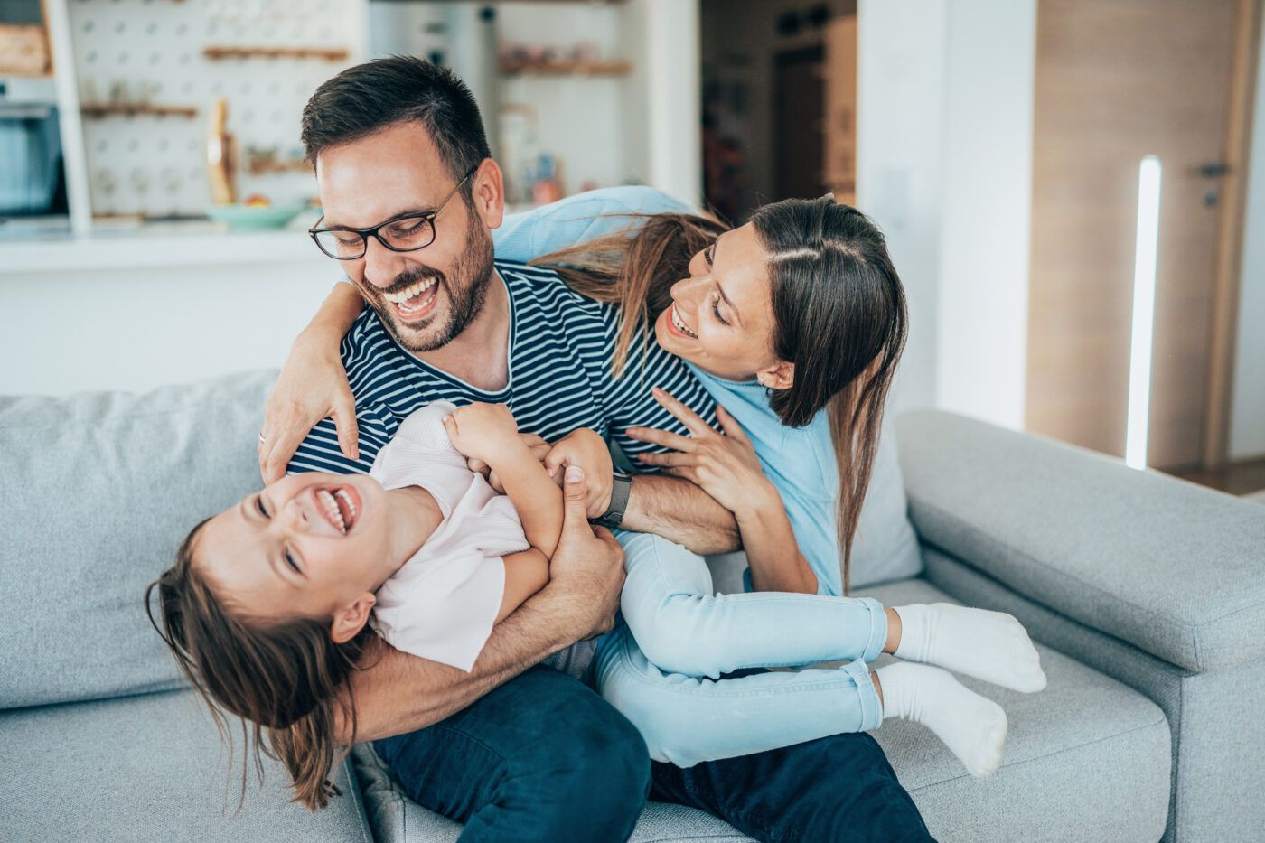 Family laughing while playfully tickling a child on a gray couch in a living room.
