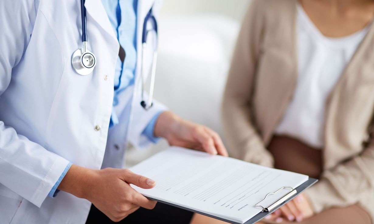 Doctor holding clipboard, facing a seated patient. Both are in a medical setting, suggesting a consultation.