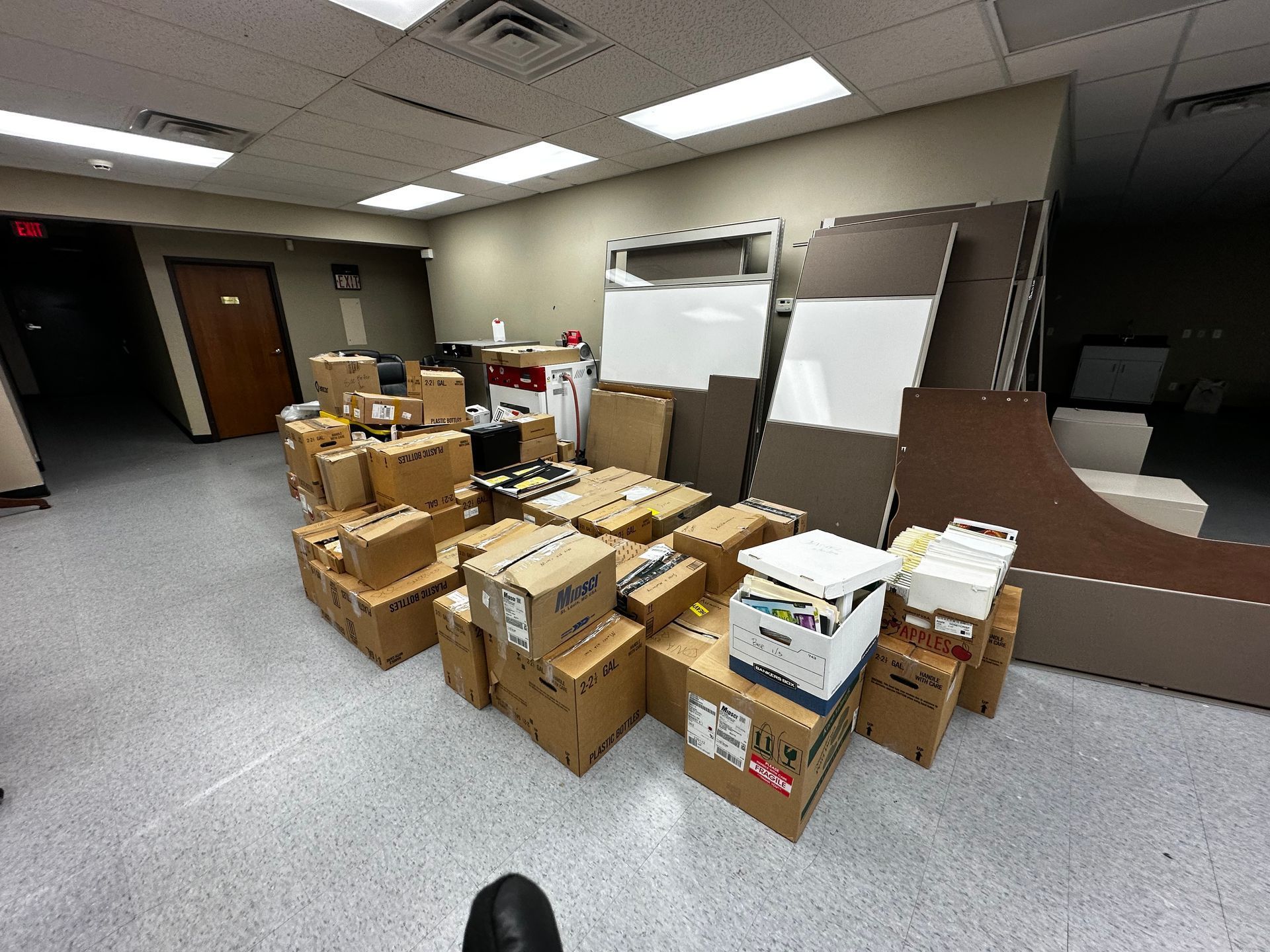 A cluttered office scene with numerous cardboard boxes piled together. The boxes vary in size and are surrounded by office furniture and supplies.