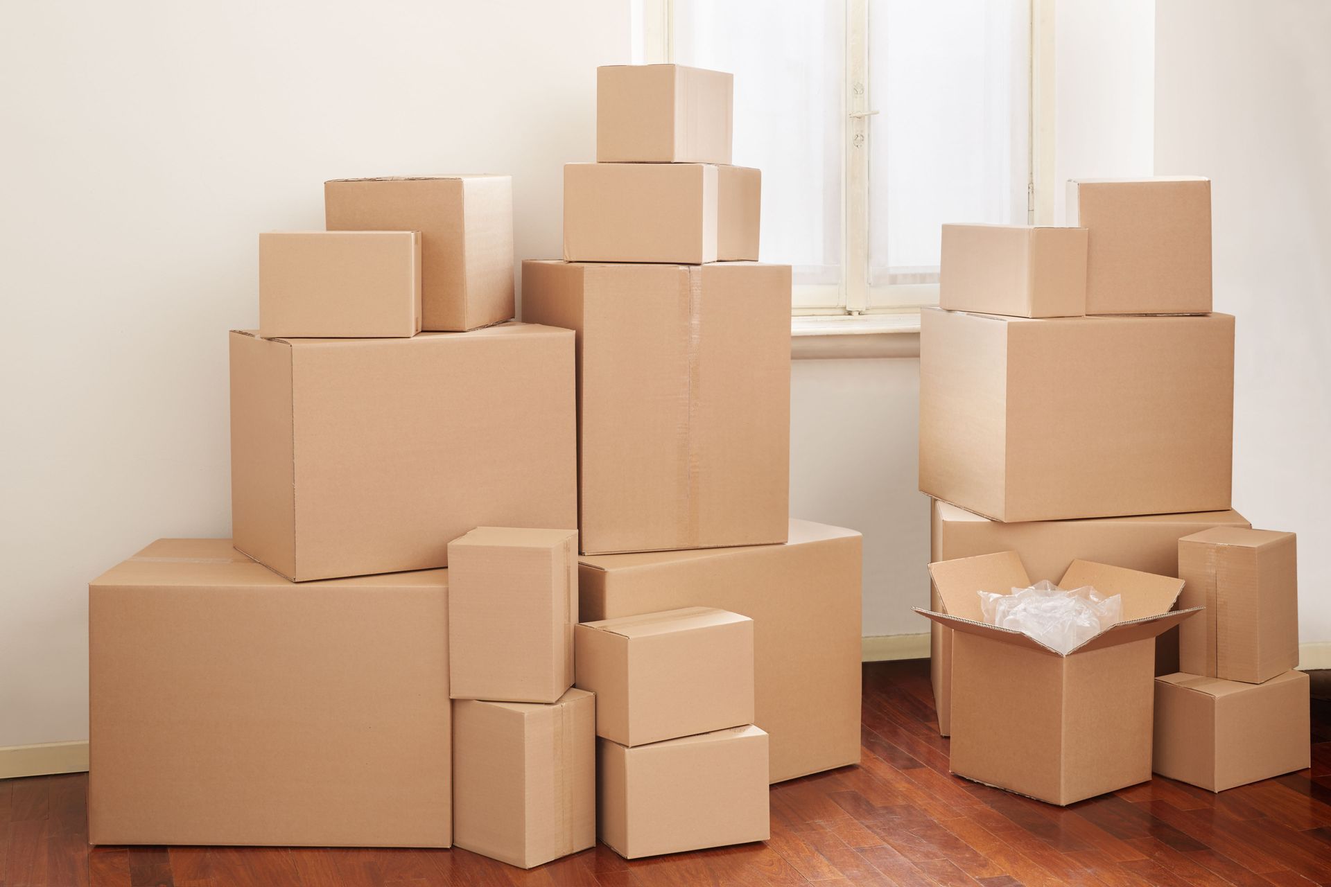 Pile of various sized cardboard moving boxes inside a room with wooden flooring, some empty, one open with packing peanuts.