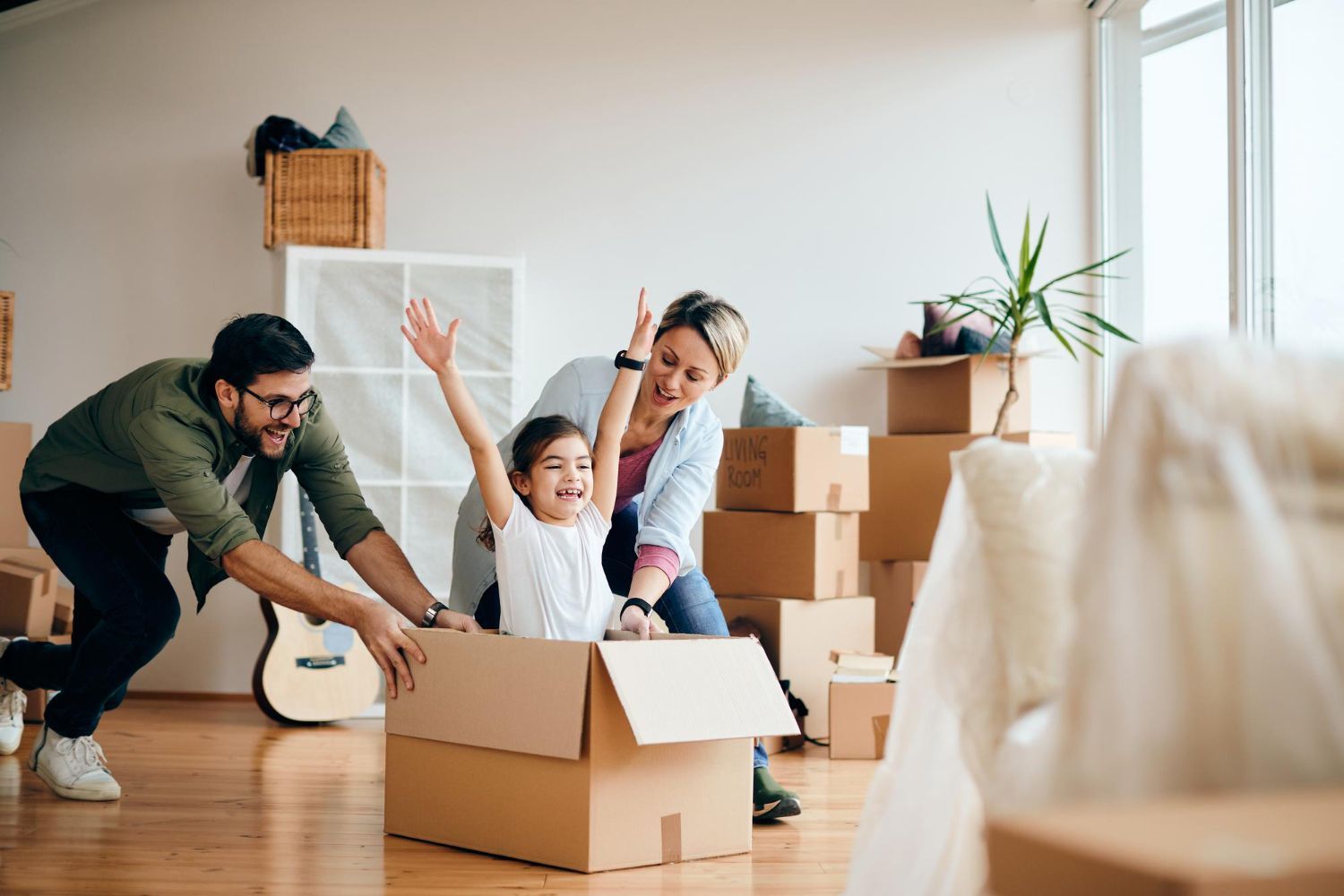 Family moving into a new home, playing with a cardboard box. Child is excitedly riding in the box, as parents push and smile.