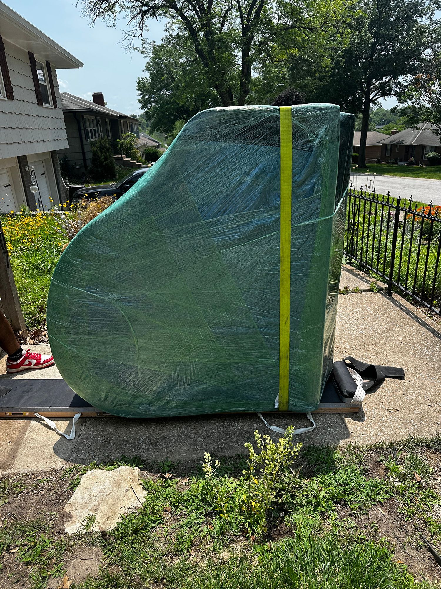 A grand piano wrapped in green plastic with a yellow strap sits on a concrete pad outside a house.