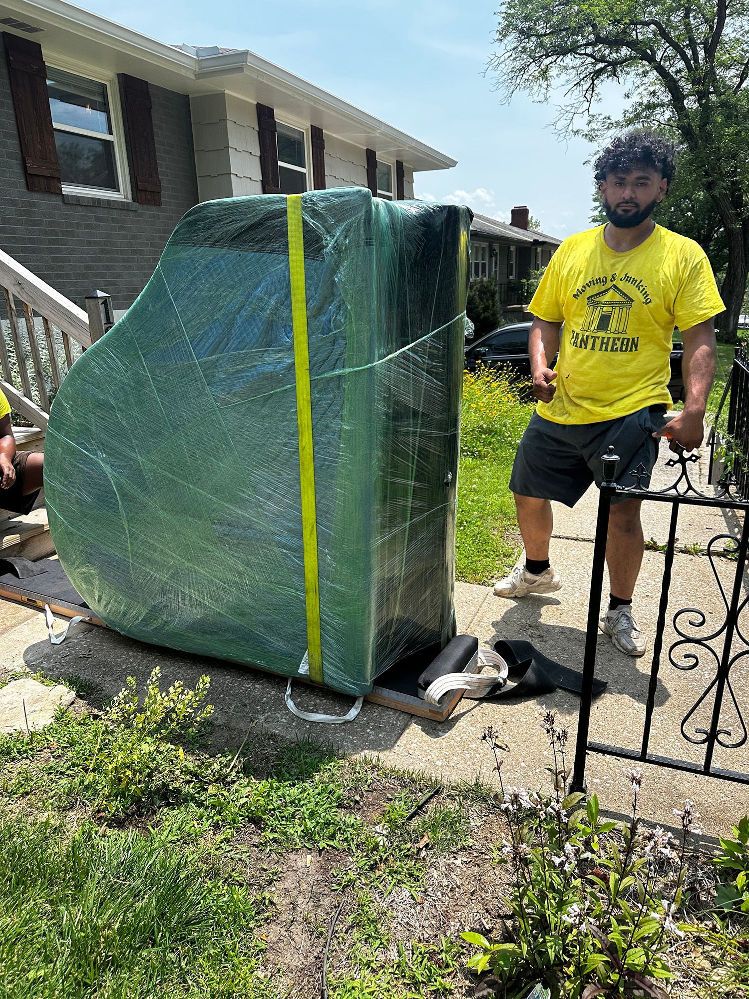 Man in yellow shirt stands next to a large piano wrapped in green plastic, ready to be moved on a sunny day.