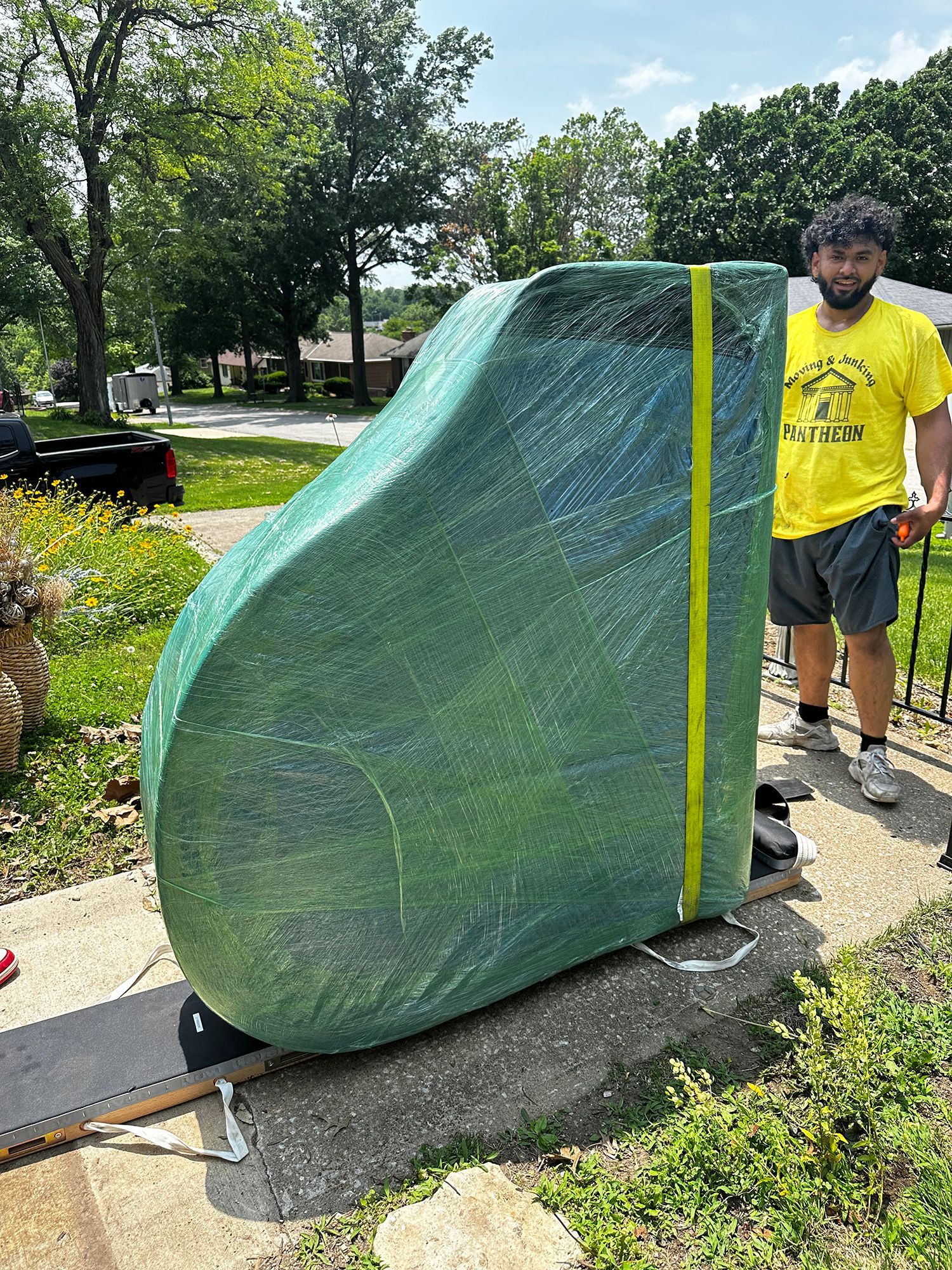 Man stands next to a wrapped, green piano being moved on a dolly in a residential setting on a sunny day.