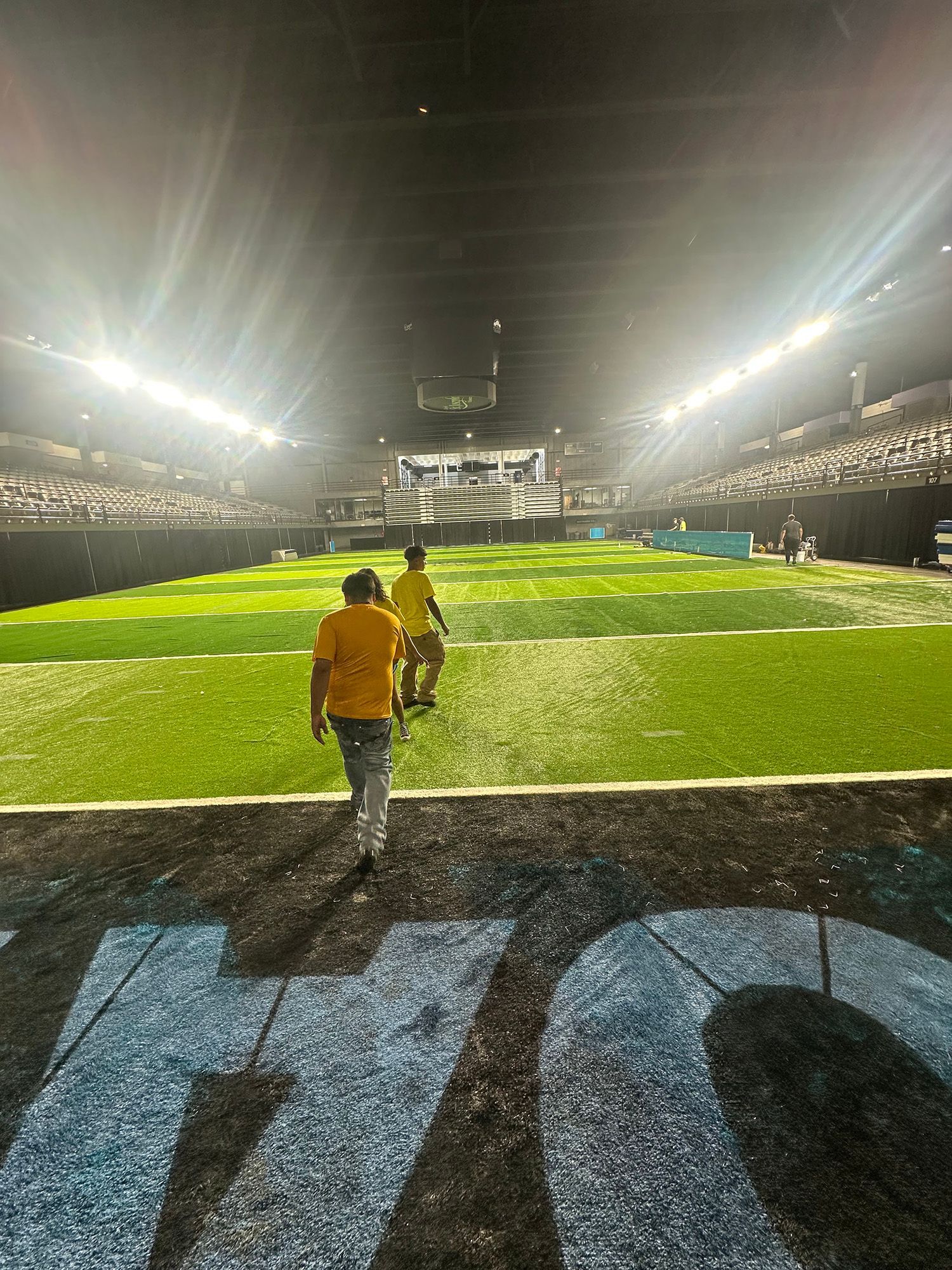 Two people walk on a football field toward the bright stadium lights. The field is green with the 40-yard line visible in the foreground.