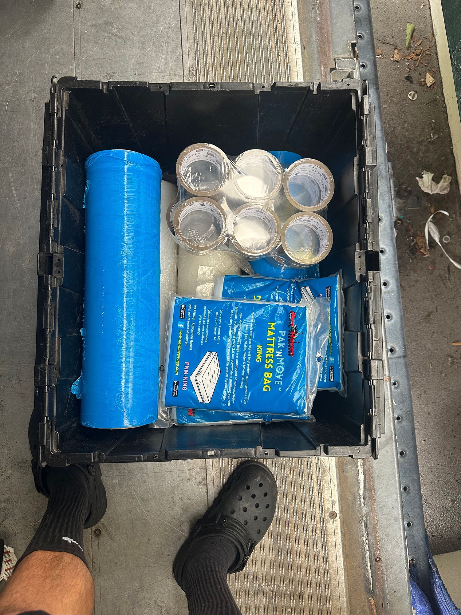 A black crate filled with blue plastic wrap, clear containers, and blue packages, viewed from above.