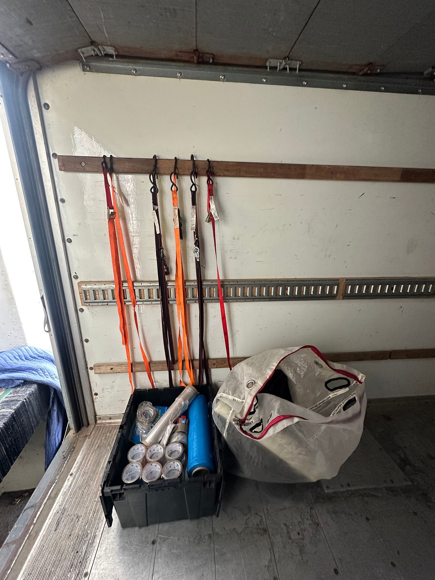 Inside a truck cargo area, a bin contains spray cans and a water bottle. Straps hang from a wall, and a trash bag rests nearby.