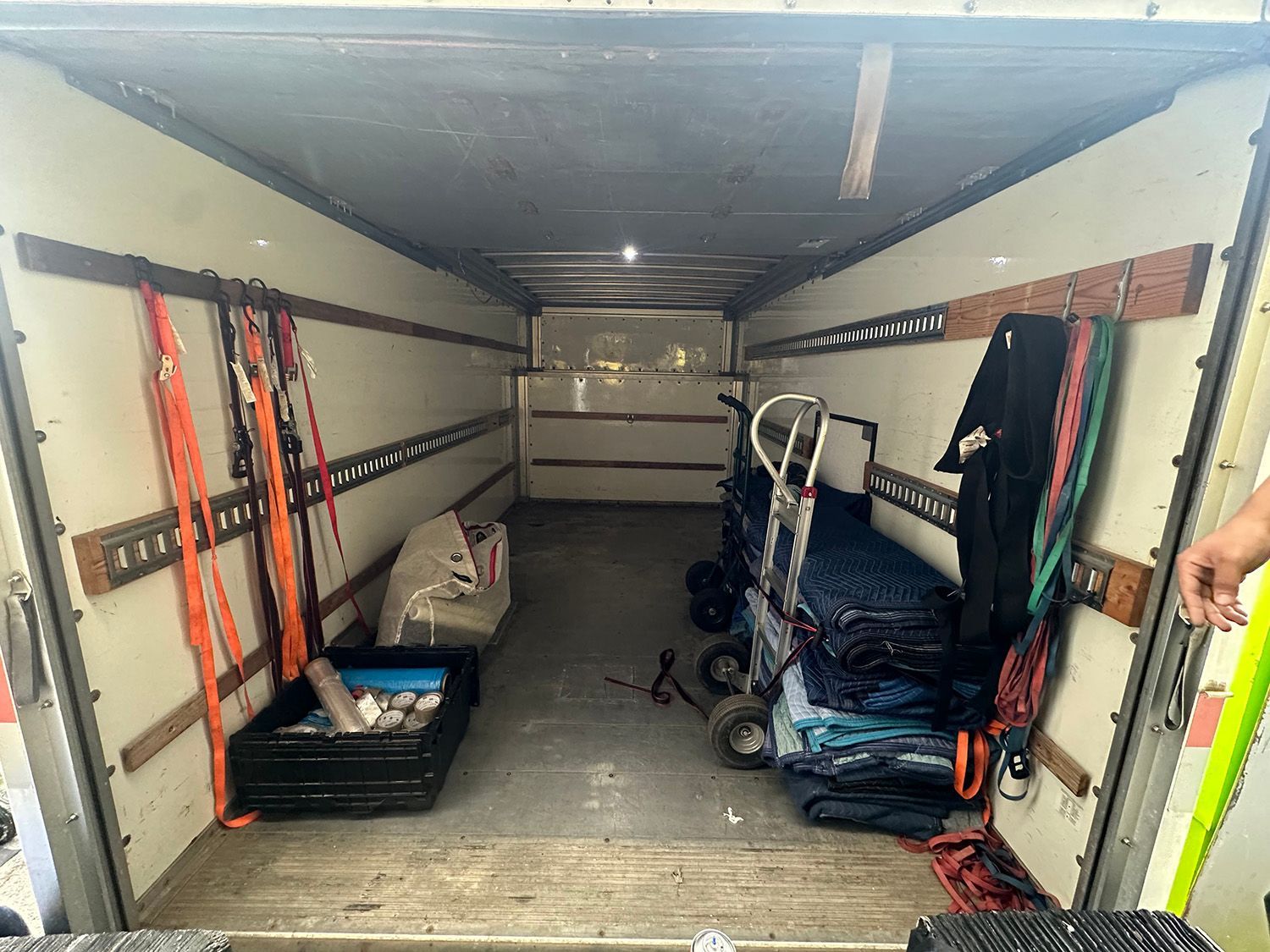 Interior view of an empty moving truck, showing straps, a dolly, and a few items. White walls and wooden rails.