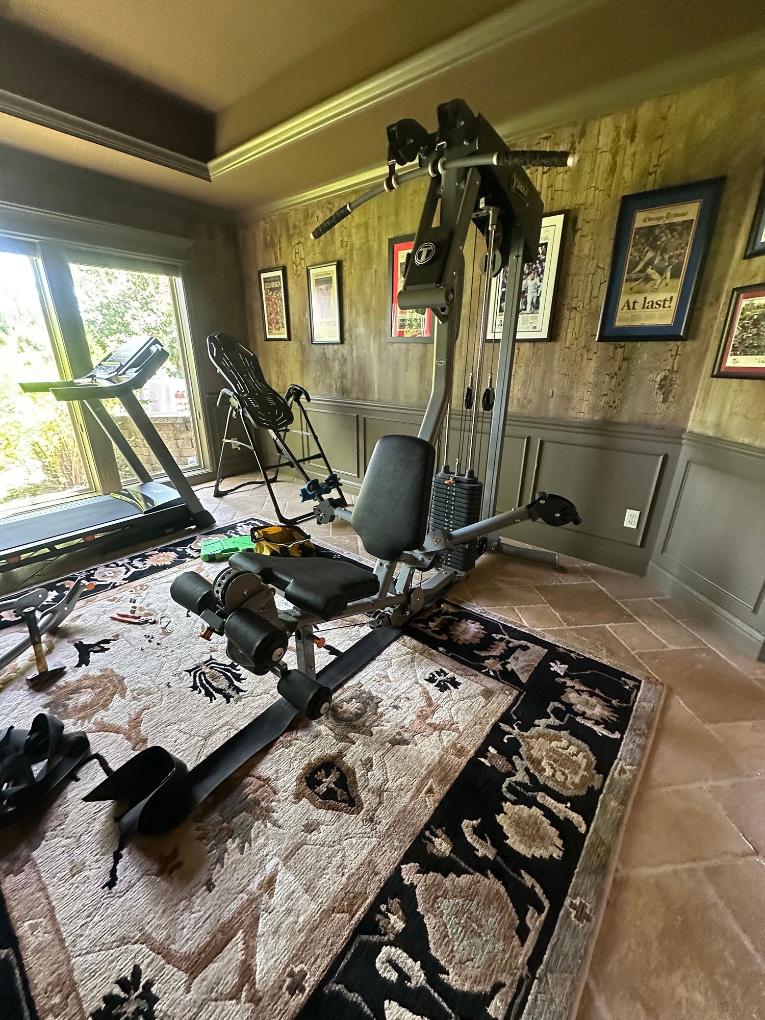 A home gym featuring a multi-station weight machine, treadmill, and inversion table on a patterned rug, against wood-paneled walls.