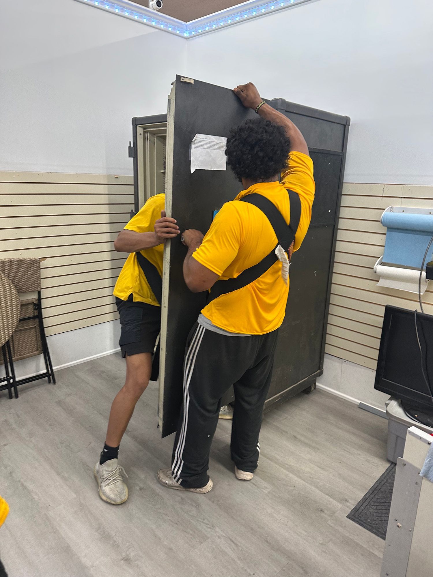 Two people wearing yellow shirts and black straps lift a large, dark panel inside a room.
