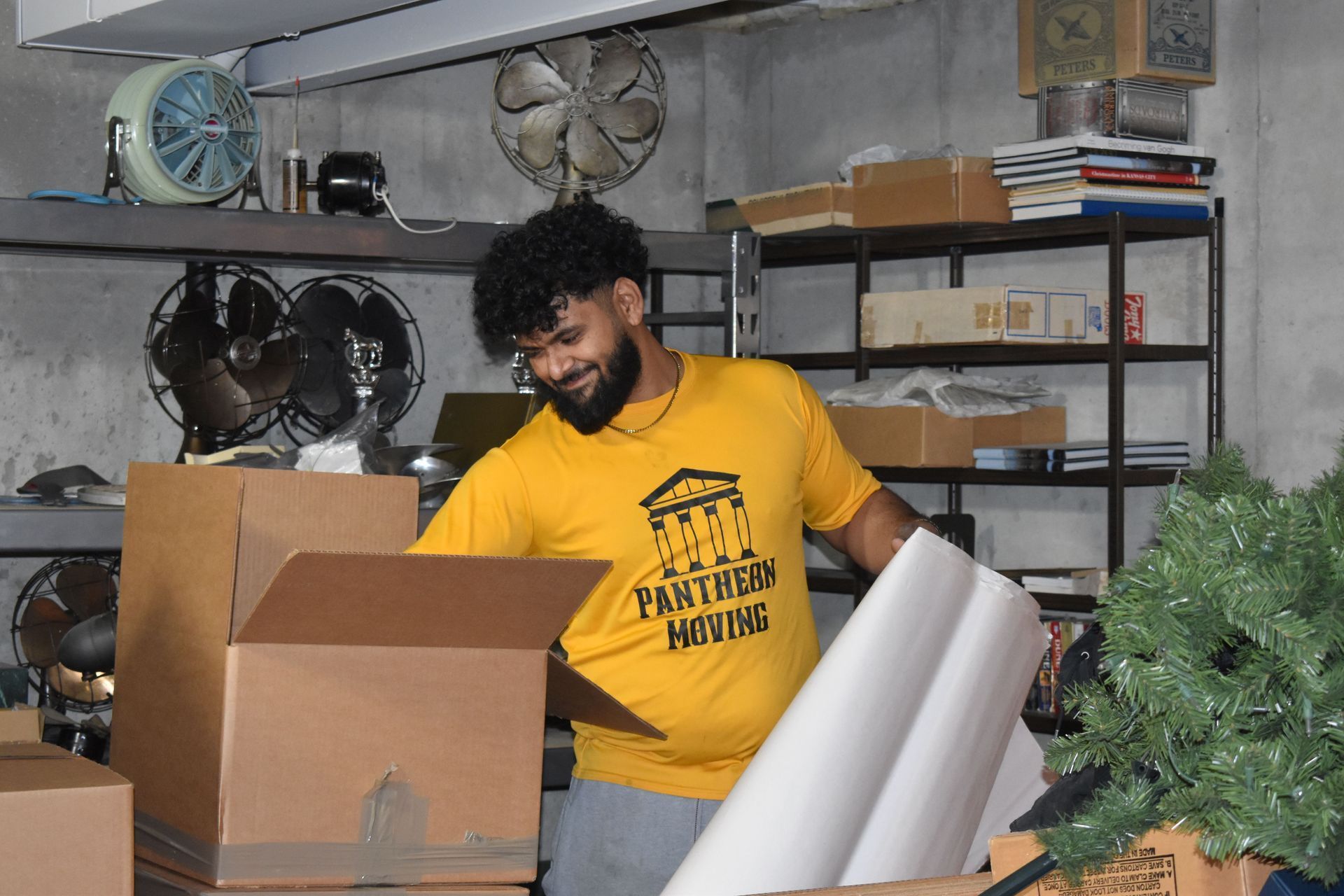 A person wearing a yellow shirt packs items into a cardboard box inside a storage room with shelves and a fake tree.