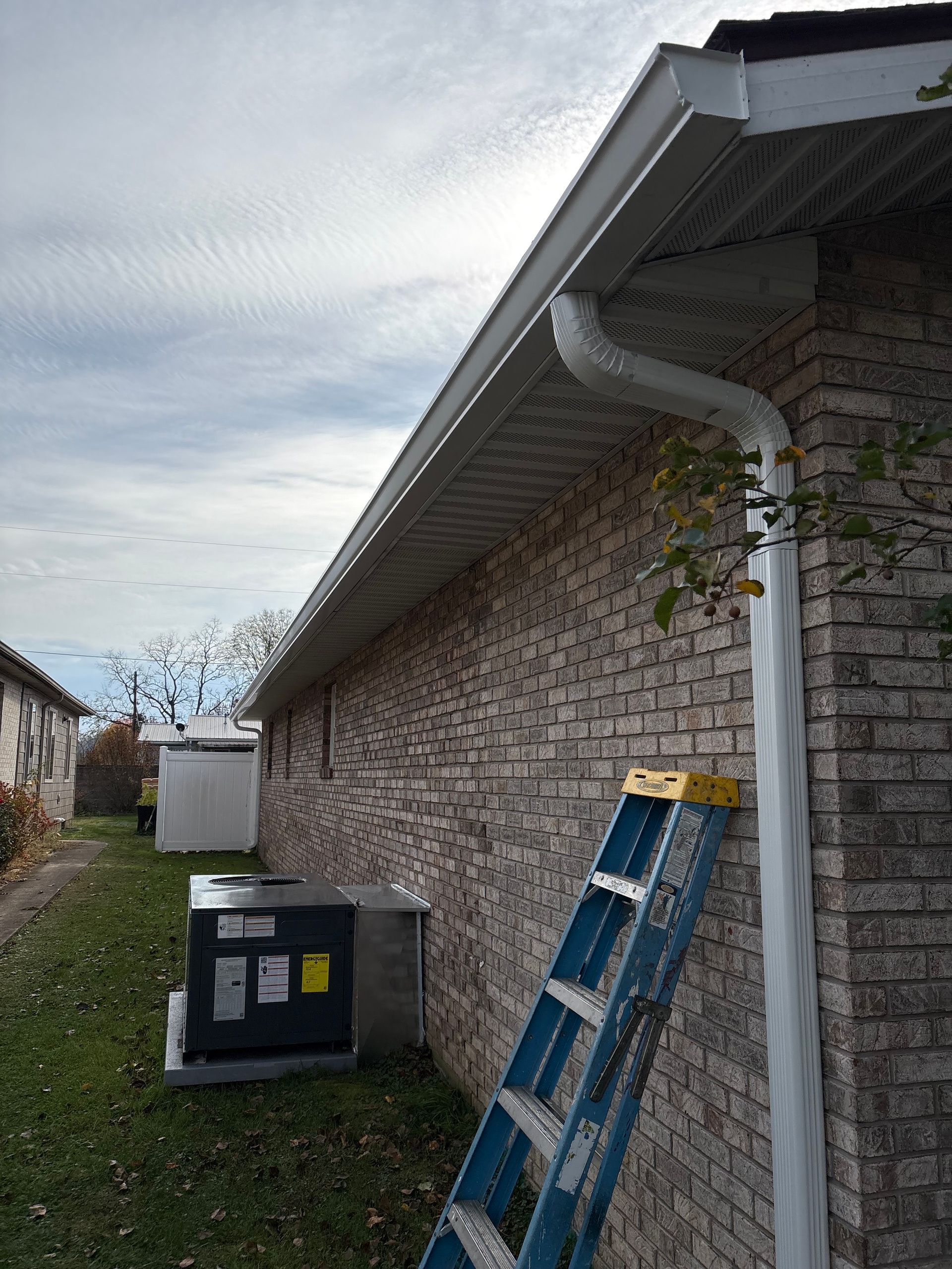 A brick building with a ladder against the side, a gutter, and an air conditioning unit on the grass.