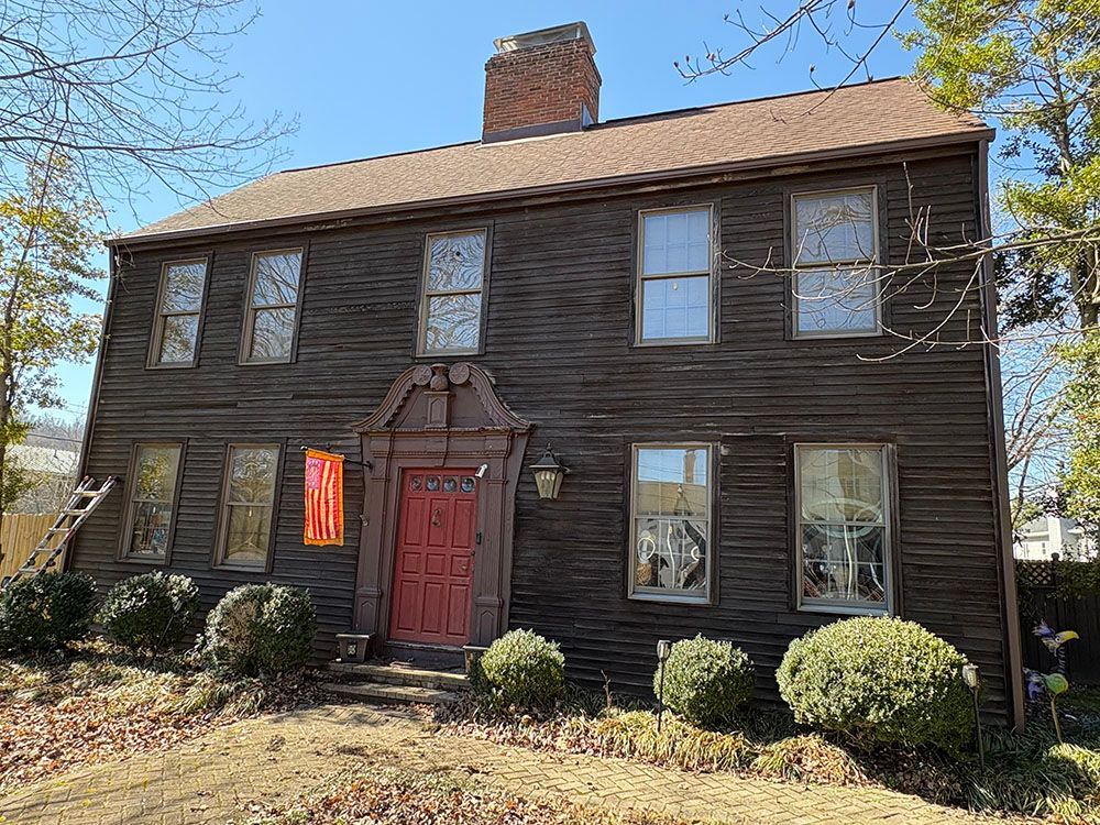 Two-story dark wood house with a red front door and brick chimney, viewed from the front yard.