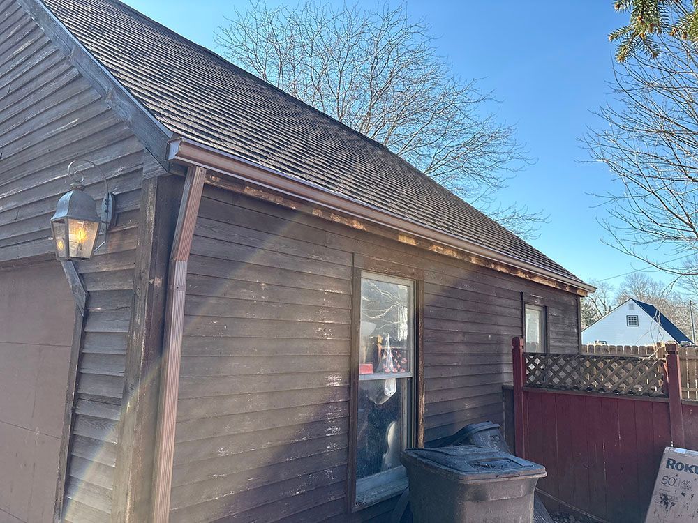 Side of a brown house with a roof, window, wall lantern, fence, and trash bins under a blue sky.