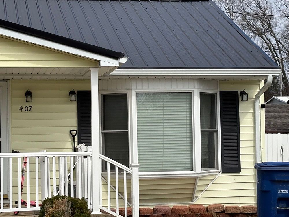 Front porch of a cream house with white railing, gray roof, and blue recycling bin.