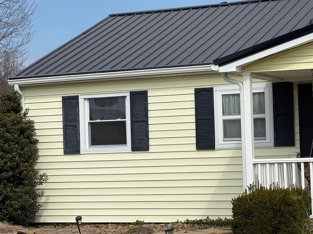 Small yellow house with a dark metal roof, black shutters, and a white porch.