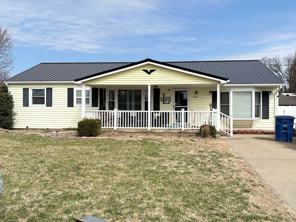 Single-story beige house with a dark roof, front porch, driveway, and a blue trash bin.