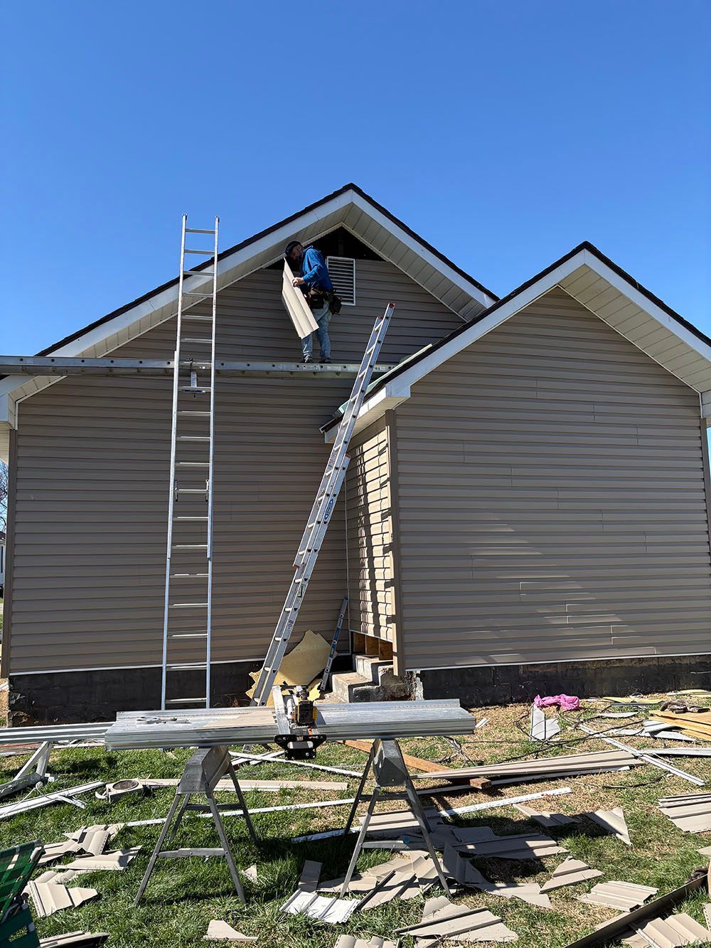 Worker on ladder repairing roof siding of a beige house on a sunny day.