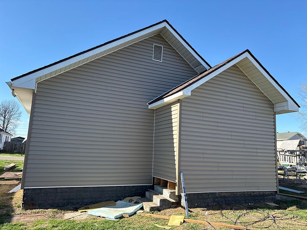 Side view of a tan house with a gabled roof and dark foundation on a sunny day.