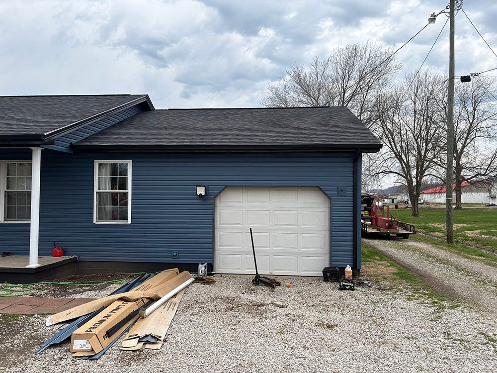 Blue house exterior with detached garage door, gravel driveway, and lumber pile under cloudy sky.