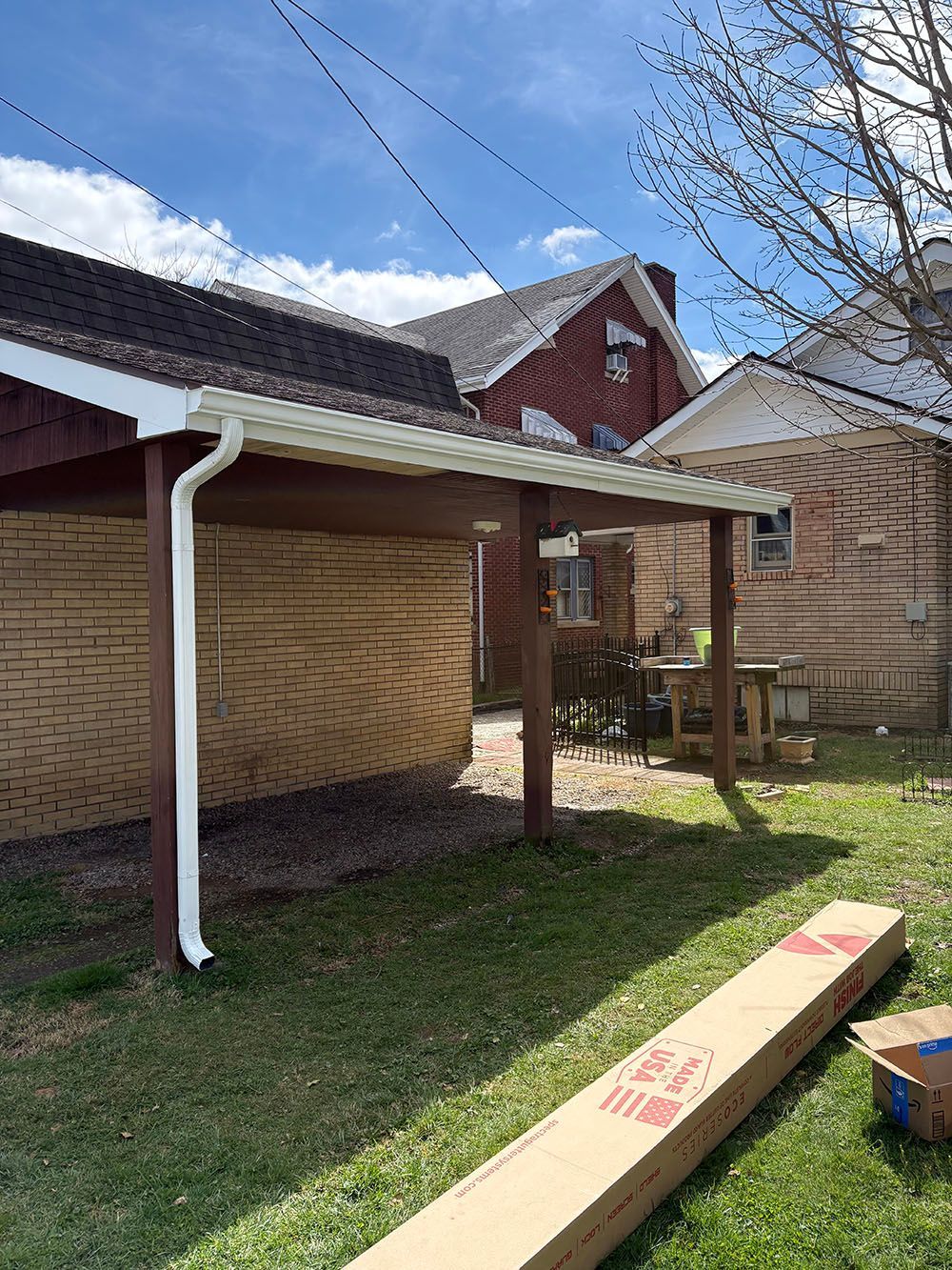 Backyard patio with brick house, wooden fence, and a long cardboard box on the grass.