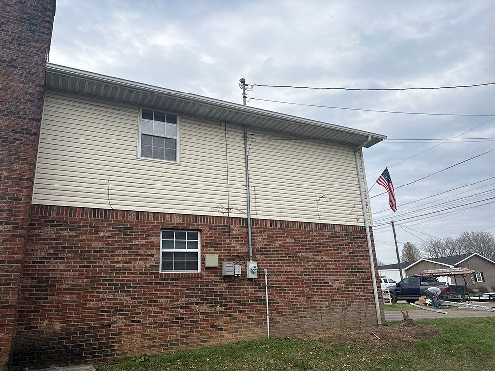 Brick apartment building with beige siding, utility meters, and an American flag by a street under cloudy skies.
