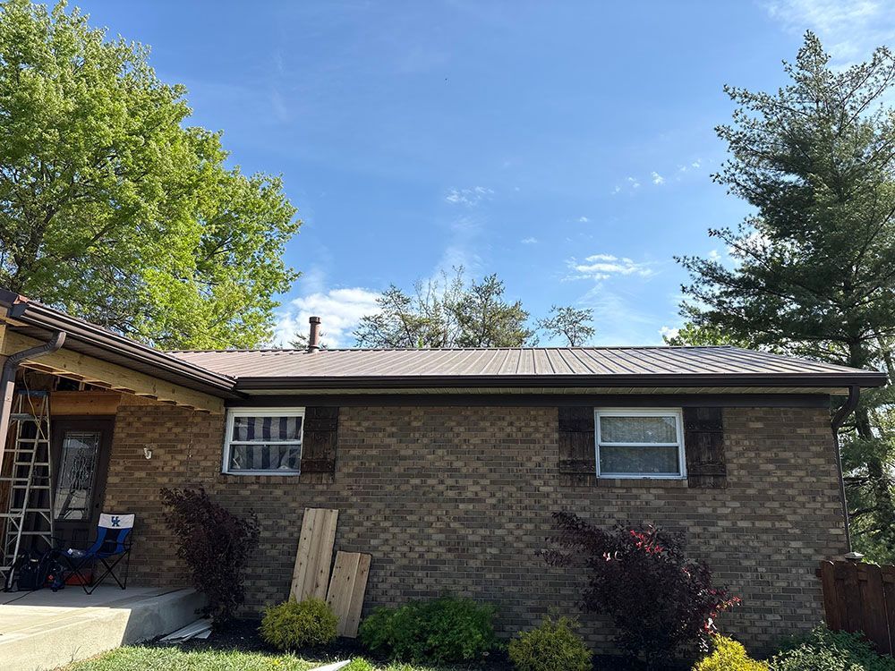 Brick house exterior with two windows, a sloped roof, and trees under a blue sky.