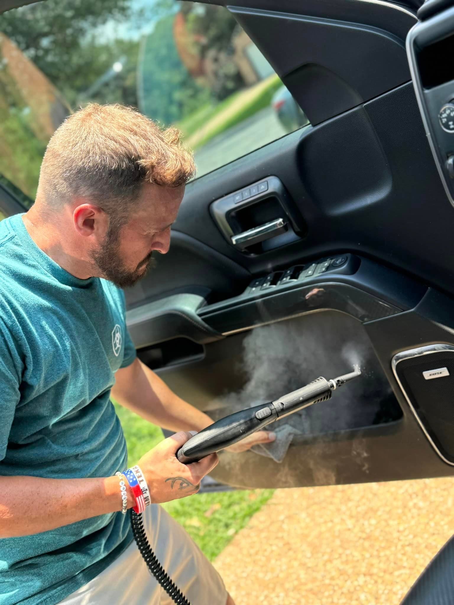 Man steaming a car door interior. He's wearing a blue shirt, outside on a sunny day.