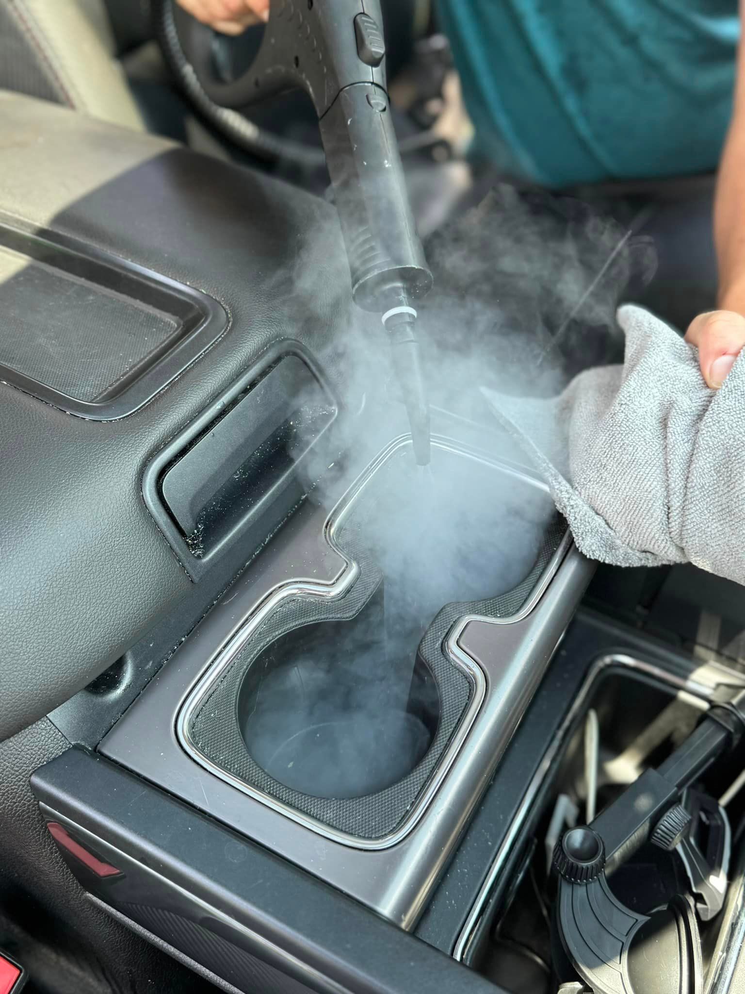 Person steam cleaning a car's center console with a handheld steamer and microfiber cloth.