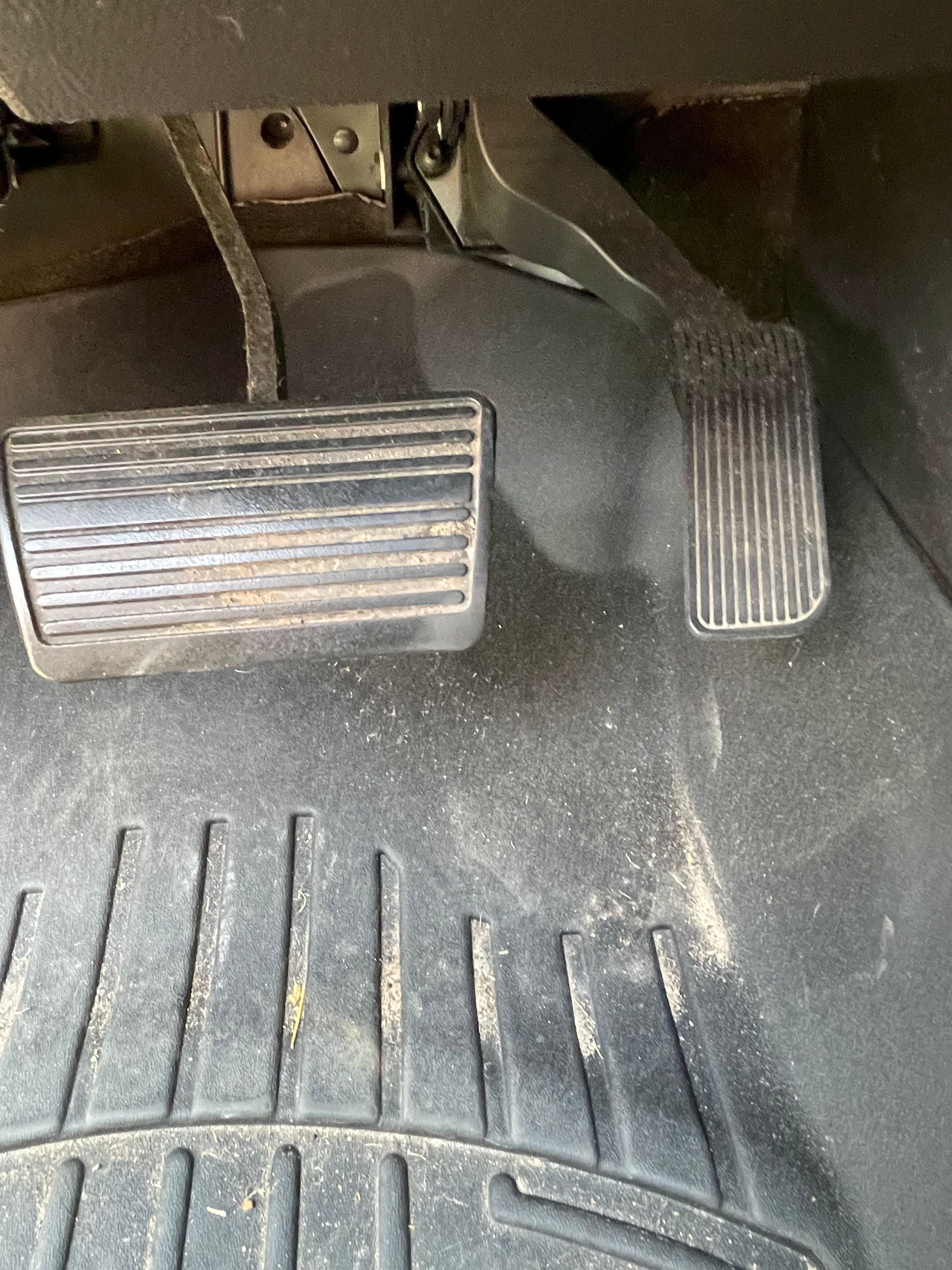 Car pedals, one larger and one smaller, on a dusty floor mat inside a vehicle.