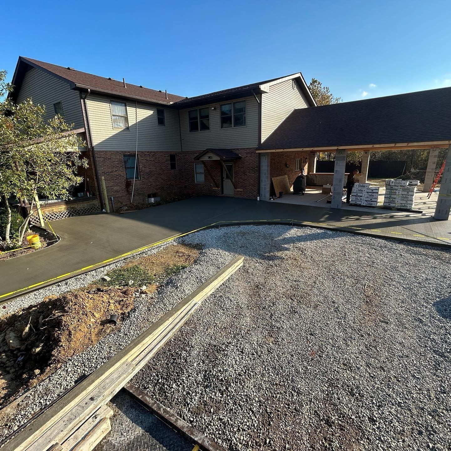 A two-story house under construction with a new concrete driveway and carport. Gravel and building materials are present.