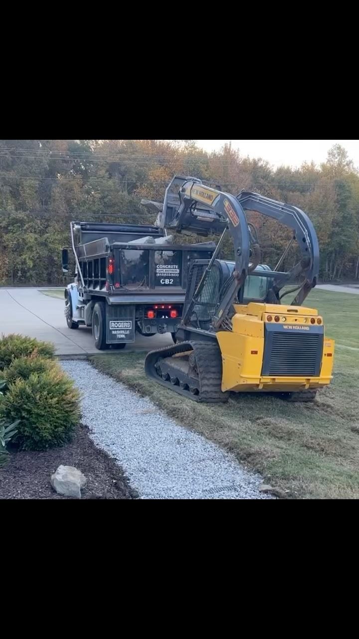 Yellow and black excavator loading a black dump truck with gravel on a paved pathway.