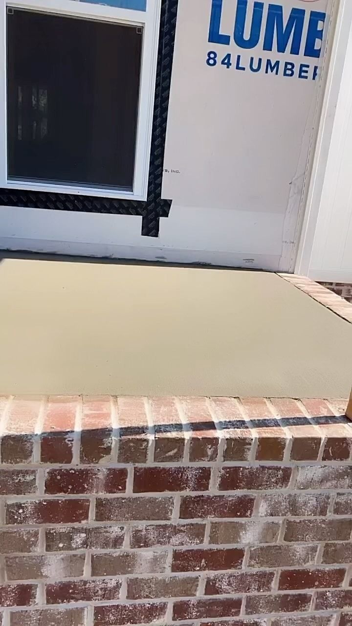 Brick-edged concrete patio with a light gray surface, below a building with a window and a 84 Lumber sign.