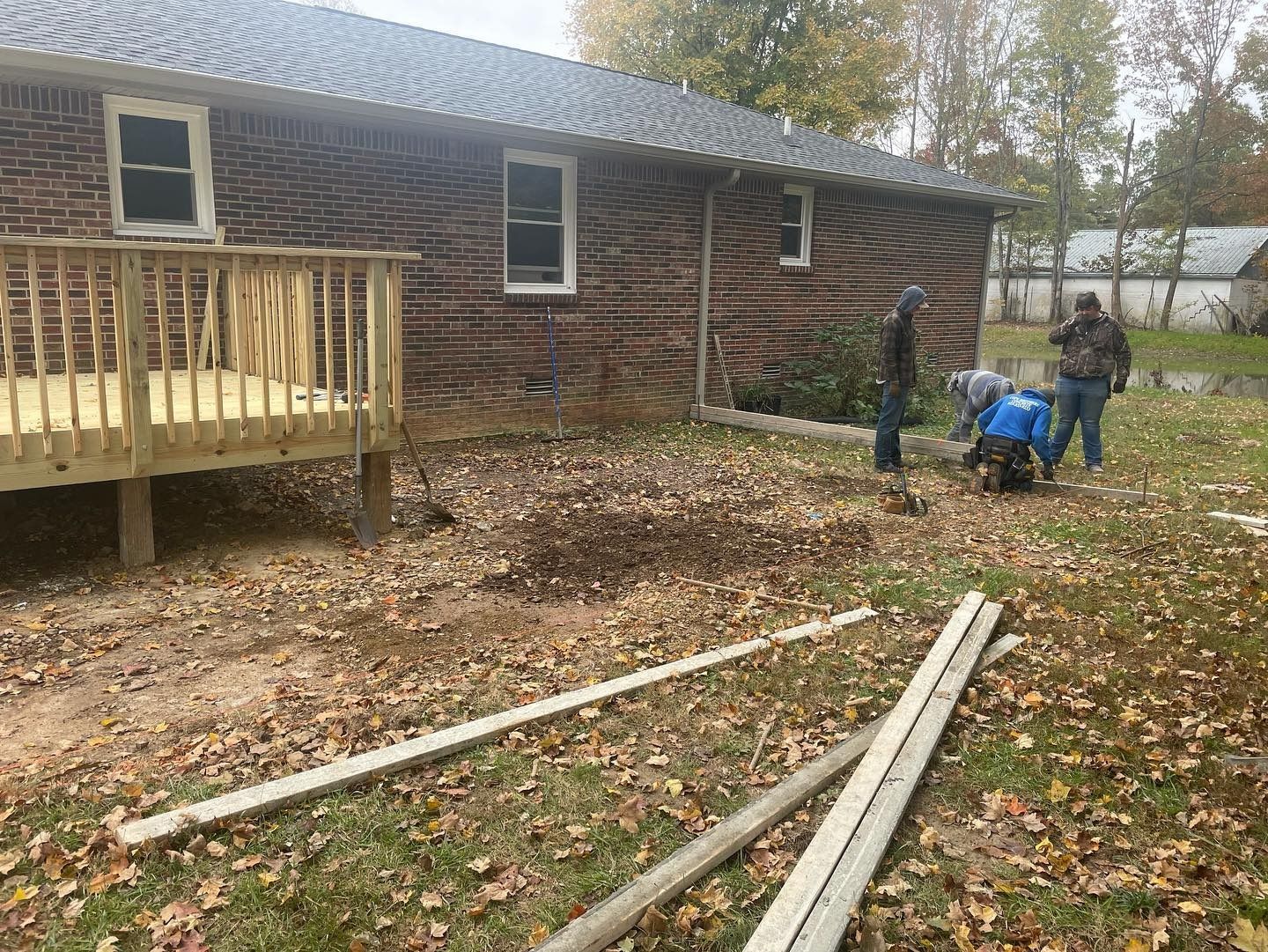 Workers preparing ground near a brick house and new wooden deck, raking leaves and debris.