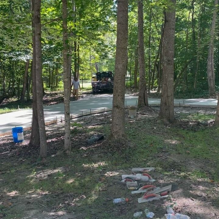 Construction of a concrete driveway in a wooded area; trees surround the wet concrete with workers in the distance.