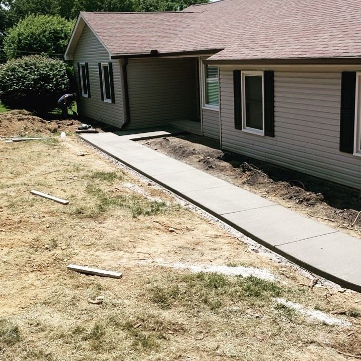 Newly poured concrete walkway leading to a house. Sidewalk borders are marked with wooden stakes; landscaping is underway.