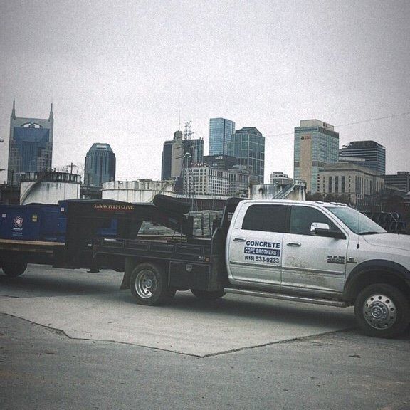 A white pickup truck with a flatbed trailer hauling materials, parked against a city skyline on an overcast day.