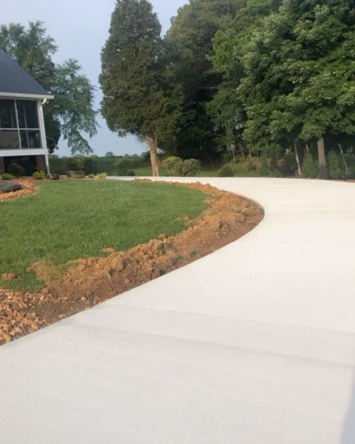 A newly paved, curved driveway winds past a grassy lawn and a house with trees in the background.
